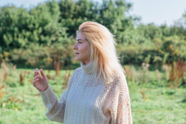 Beautiful blond woman staying and smocking cigarette in a green field - Фото, зображення