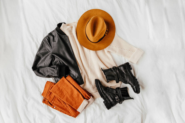 Leather jacket and white warm sweater with brown trousers and hat on white sheet. Women's stylish autumn or winter outfit. Trendy clothes collage. Flat lay, top view.  - Fotografie, Obrázek