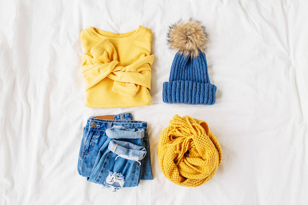 Cozy yellow sweater, blue jeans, scarf and hat on bed on white sheet. Women's stylish autumn or winter outfit. Trendy  clothes collage. Flat lay, top view.  - Fotografie, Obrázek
