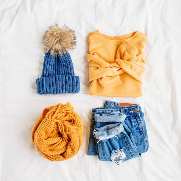 Cozy yellow sweater, blue jeans, scarf and hat on bed on white sheet. Women's stylish autumn or winter outfit. Trendy  clothes collage. Flat lay, top view.  - Fotografie, Obrázek