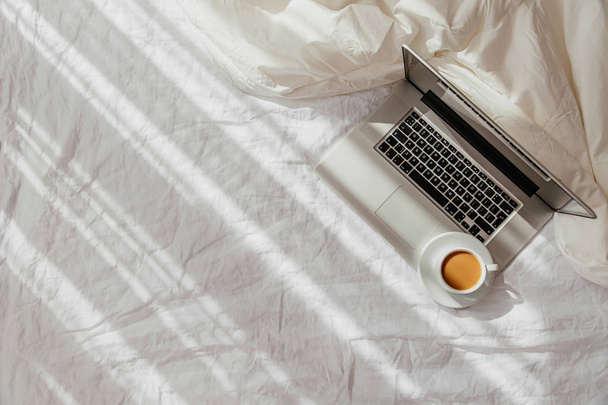 Laptop and cup of coffee on white bed with a  blanket. Work at home concept. Morning light  - Foto, imagen