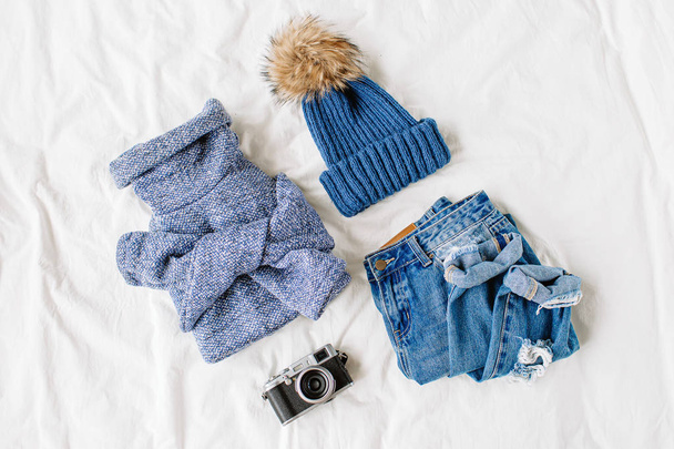 Cozy blue sweater,  jeans,  hat and photo camera on bed on white sheet. Women's stylish autumn or winter outfit. Trendy  clothes collage. Flat lay, top view.  - Fotografie, Obrázek