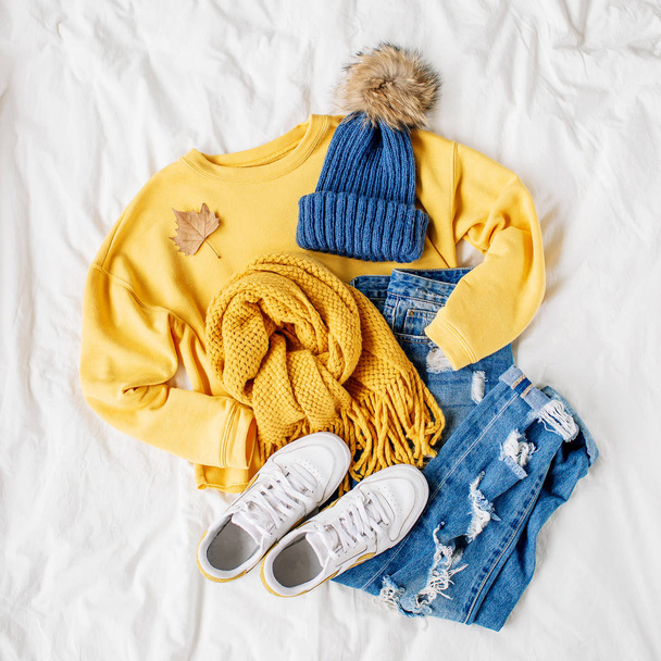 Cozy yellow sweater, blue jeans, sneakers, scarf lying on bed on white sheet. Women's stylish autumn or winter outfit. Trendy  clothes collage. Flat lay, top view.  - Fotografie, Obrázek