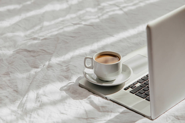 Laptop and cup of coffee on white bed. Work at home concept. Morning light  - Fotografie, Obrázek
