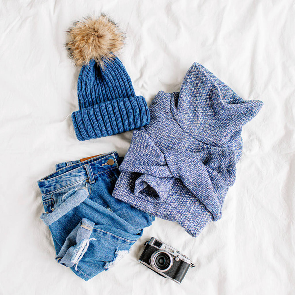 Cozy blue sweater,  jeans,  hat and photo camera on bed on white sheet. Women's stylish autumn or winter outfit. Trendy  clothes collage. Flat lay, top view.  - Fotografie, Obrázek
