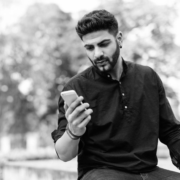Jeune bel homme indien utilisant un téléphone portable tout en étant assis sur le sol en béton au parc
 - Photo, image