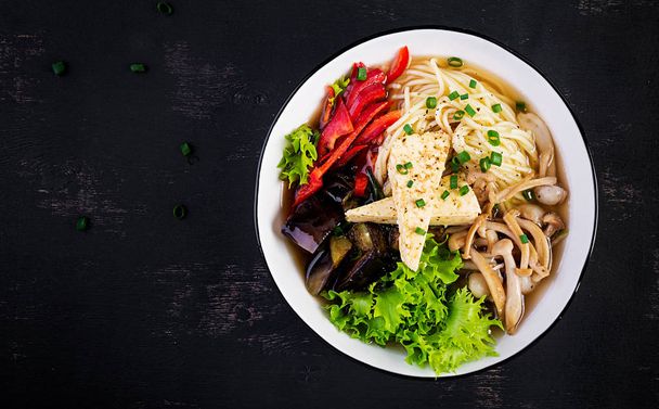 Asian vegan noodles soup with tofu cheese, shimeji mushrooms and fried eggplant in bowl on dark background. Top view - Photo, Image