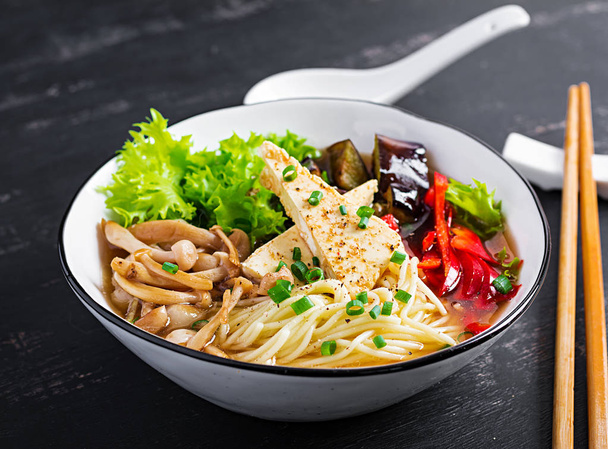 Asian vegan noodles soup with tofu cheese, shimeji mushrooms and fried eggplant in bowl on dark background. - Photo, Image