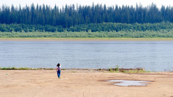 Une touriste avec un sac à dos touchant ses cheveux, solitaire va à la falaise de béton de la rive sur la vieille jetée à la rivière Nord vilyu avec la toundra d'épinette de la forêt de Yakutia de l'autre côté
. - Photo, image