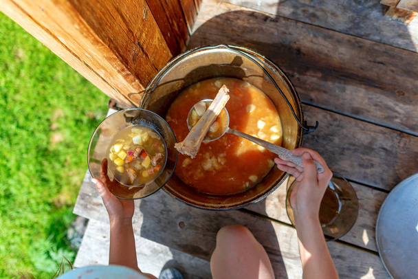 Chica chef sosteniendo cuchara en el hueso y repartiendo sopa en un tazón transparente sopa de la caldera con una comida al aire libre en el suelo de madera en el bosque
. - Foto, Imagen