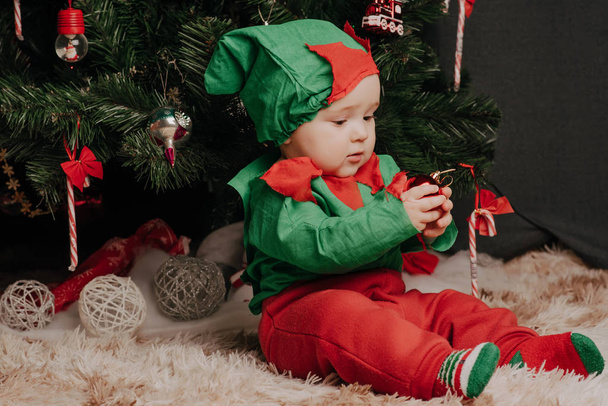 маленький хлопчик в ельфійському костюмі сидить під ялинкою з кульками
 - Фото, зображення