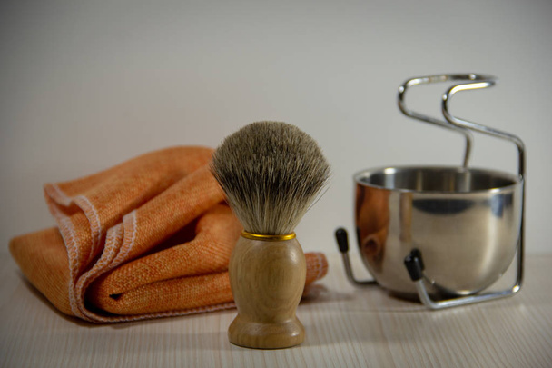 Accesorios de afeitado: una afeitadora de madera con un cepillo de cerdas naturales, un recipiente de metal en un soporte, una toalla naranja. Fondo claro
. - Foto, imagen