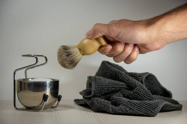 Ένα αρσενικό χέρι φέρνει ένα ξύλινο ξυράφι ξυρίσματος με μια βούρτσα σε ένα μεταλλικό μπολ με αφρό σε μια βάση, δίπλα σε μια γκρίζα πετσέτα. Αξεσουάρ ξυρίσματος. Φως φόντο. - Φωτογραφία, εικόνα