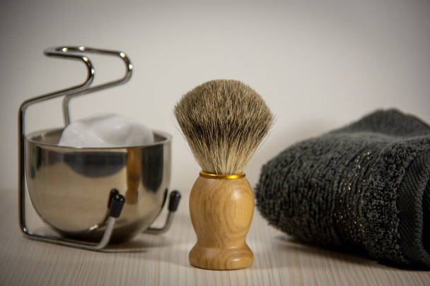 Accesorios de afeitado: una afeitadora de madera con un cepillo, un recipiente de metal con espuma en un soporte, una toalla gris. Fondo claro
. - Foto, imagen