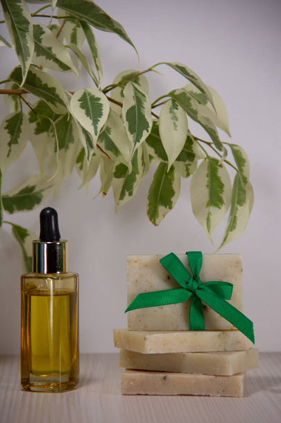 緑の弓で飾られた手作りの石鹸、ドロッパー付きのガラス瓶の中の黄金の顔油、薄い背景に葉を持つベンジャミンの枝. - 写真・画像