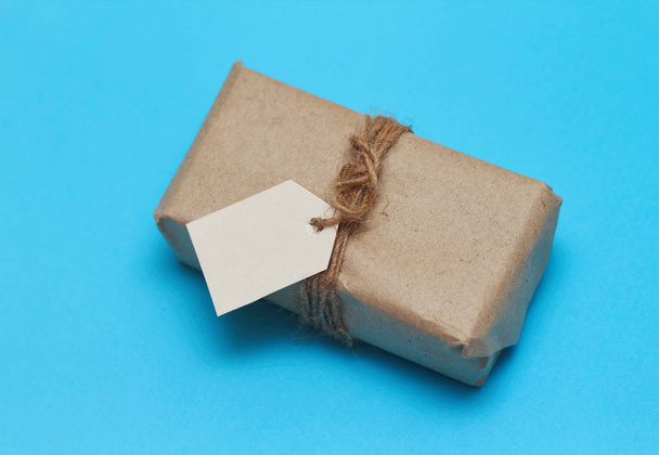 Parcel wrapping in brown craft paper and tie hemp string. Package with paper label. Delivery service. Online shopping. Your purchase. Gift box on a table. Blue solid background. - Photo, image