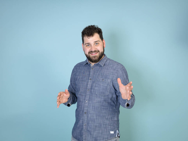 cool and young man with black hair and black beard grimacing while posing in front of blue wall background   - 写真・画像