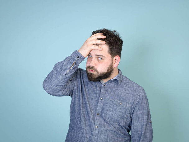 Frowning man with black hair and black beard holding his painful head and looking up while posing in front of blue wall background - Foto, Imagem