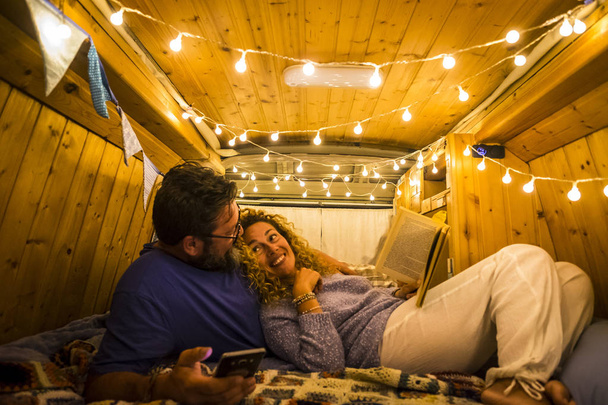 δύο ενήλικες που ζουν σε ένα όμορφο και όμορφο φορτηγάκι ξαπλωμένοι στο κρεβάτι διαβάζοντας ένα βιβλίο και χρησιμοποιώντας το τηλέφωνο  - Φωτογραφία, εικόνα