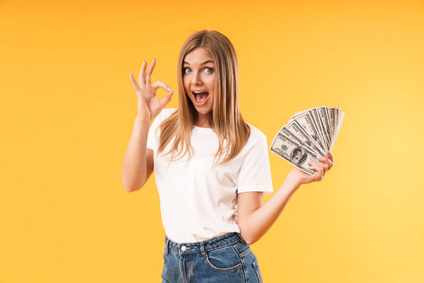 Imagen de primer plano de la mujer rubia feliz con camiseta casual que muestra signo de ok mientras sostiene el dinero en efectivo
 - Foto, Imagen