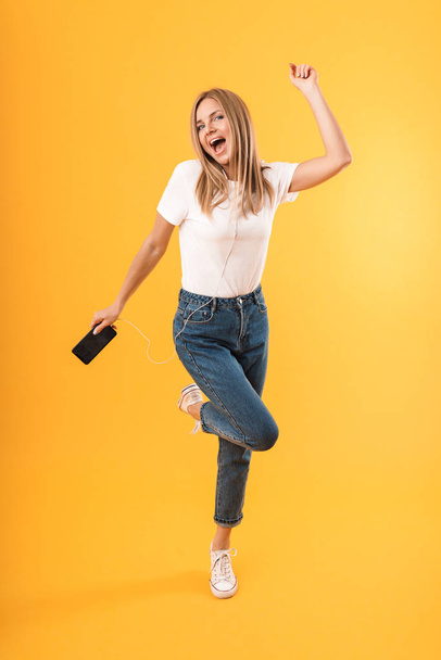 Full length image of cheerful blond woman smiling while listening to music with smartphone and earphones - Foto, afbeelding