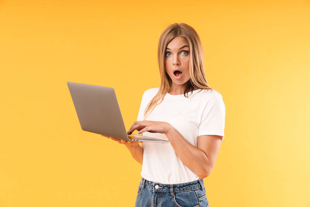 Image of perplexed blond woman wearing casual t-shirt expressing wonder while using laptop computer - 写真・画像