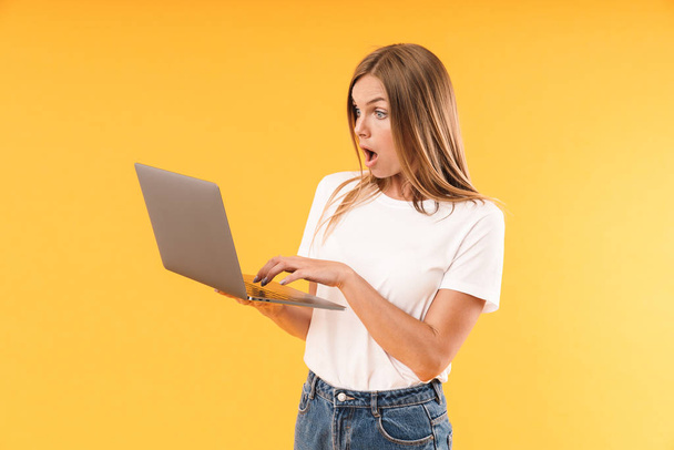 Image of surprsied blond woman wearing casual t-shirt expressing wonder while using laptop computer - Foto, Imagem