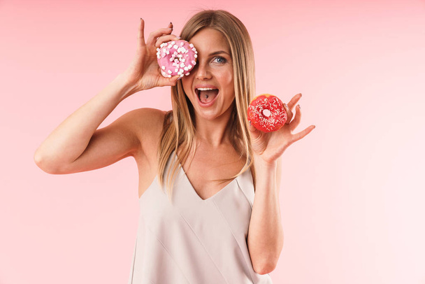 Image of joyous blond woman wearing dress smiling while holding two sweet donuts - Foto, imagen