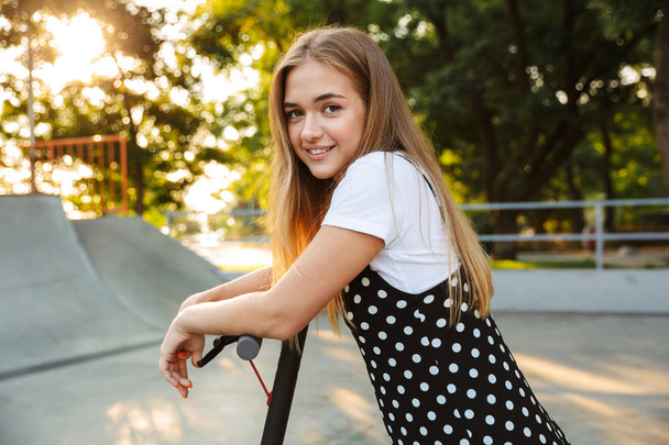 Optimistic cheerful teenage girl in park walking on scooter. - Fotó, kép