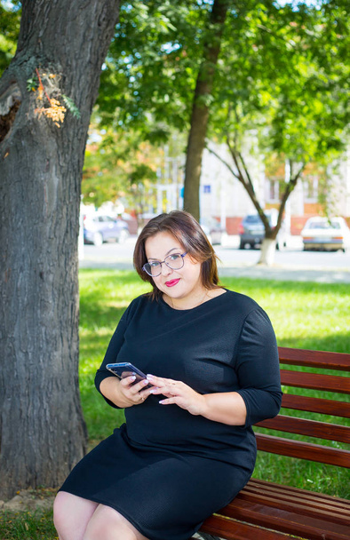 Красивая пухленькая девушка в черном платье сидит на скамейке в парке с телефоном в руке
 - Фото, изображение