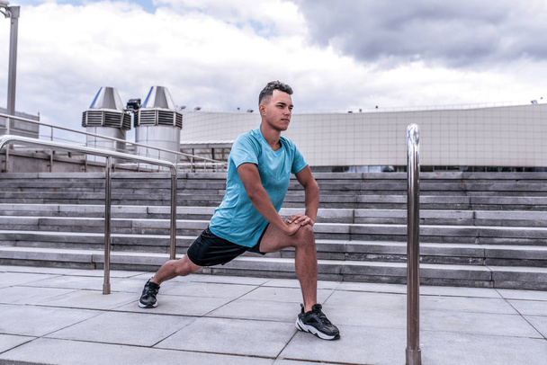 Male athlete guy, in summer in city, doing warm-ups of knees of joints and legs, ankle-boot, before morning jogging, fitness workout training, background concrete steps of cloud. - Φωτογραφία, εικόνα