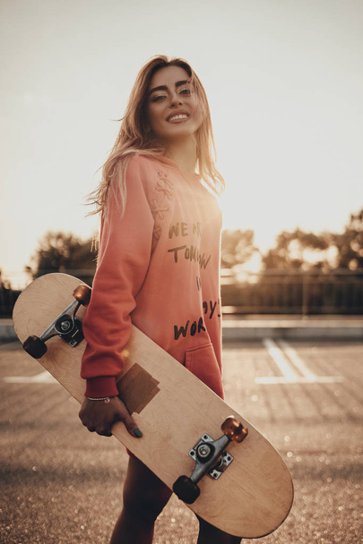 Happy stylish girl with skateboard in red hoodie on city street. Positive fashion young woman outdoors portrait with longboard in sunset evening - Фото, изображение