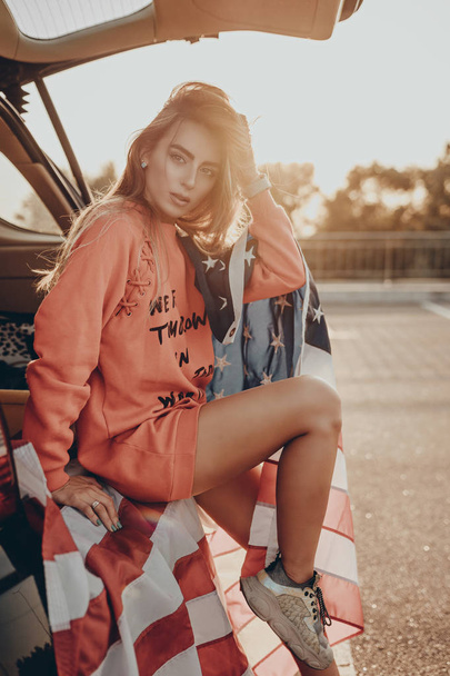 Atrakcyjny, stylowy nastolatek siedzący w samochodzie na parkingu z amerykańską flagą w czerwonej bluzie i tenisówkach. Stylowa moda młoda aktywna dziewczyna pozowanie w zachód słońca w lecie na ulicy miasta - Zdjęcie, obraz