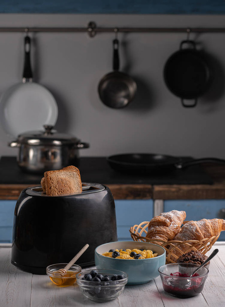 Comida casera deliciosa. Tostadas frescas, croissant dulce, copos de maíz con bayas y miel en la mesa blanca en la cocina
 - Foto, Imagen