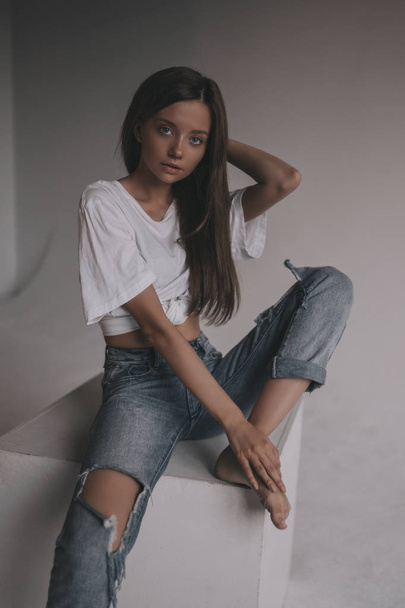 Beautiful stylish woman in shirt, jeans and sneakers posing in studio on cube on white background. Seductive glamour young girl model portrait. - 写真・画像