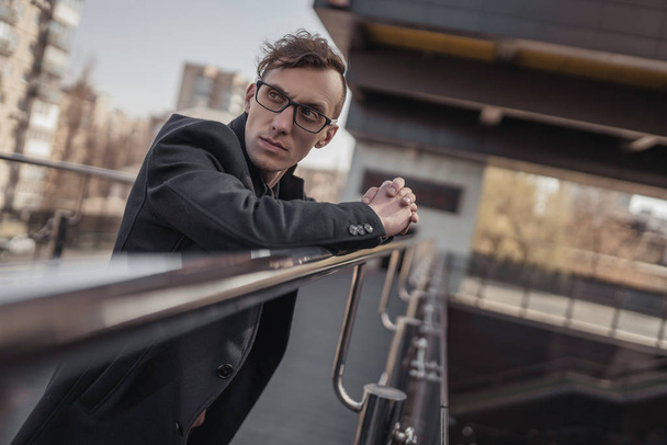 Portrait of stylish handsome man on the bridge. Fashion shooting on the city street. Wearing in black coat and glasses. Confident looking - 写真・画像