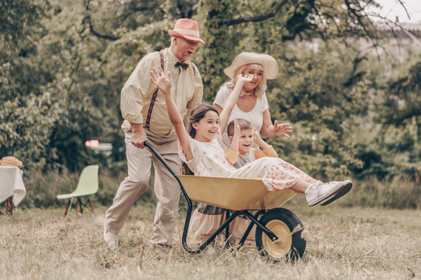 Бабушка с дедушкой в парке с внуками, играющими с телегой. Семья веселится в саду. Старики рады видеть внучку и внука
 - Фото, изображение