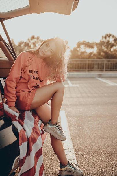 Boldog elegáns lány amerikai zászlóval piros kapucnis pulcsiban a város utcáján. Gyönyörű divat fiatal nő szabadban portré autóban parkolás naplemente este - Fotó, kép