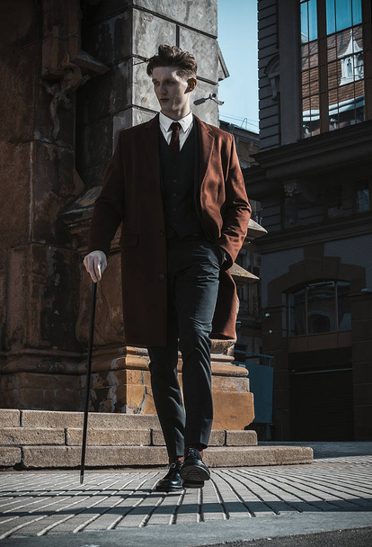 Fashion portrait of young man on red coat, white shirt, black suit and cane walking on streets of city background. Model shooting - Photo, image