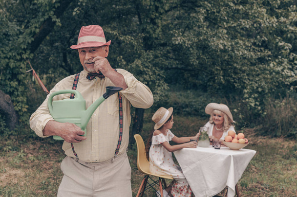 Ein glücklicher Großvater mit einer Gießkanne zwinkert und blickt in die Kamera. bekleidet mit Hemd, Hose, Fliege und Hut. Im Hintergrund eine Großmutter mit ihrer Enkelin, die Tee trinkt und Obst isst - Foto, Bild