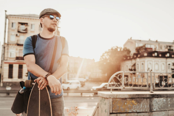 Ευτυχισμένος άνθρωπος με γυαλιά ηλίου, μπλε πουκάμισο και καπέλο που θέτουν με skateboard κοντά στο δρόμο στο παρασκήνιο. Εξωτερικό πορτρέτο του στιλάτου άντρα. Αστικός τρόπος ζωής - Φωτογραφία, εικόνα