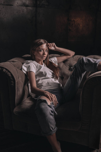 sexy heiße Frau in Hemd und Jeans posiert in Loft-Studio auf Sessel auf schwarzem Hintergrund. verführerischer Glamour junges Mädchen Modell Porträt. - Foto, Bild