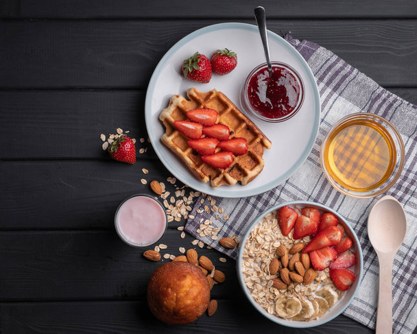 Healthy food. Breakfast. Granola with milk, fruit and nuts. Delicious dessert - crispy belgian waffle with strawberries and jam. Honey, yogurt, chocolate muffin. On a black background. Top view - Фото, изображение