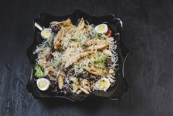 Вкусный свежий салат Цезарь с курицей и яйцами в черной тарелке на фоне черного каменного стола
 - Фото, изображение