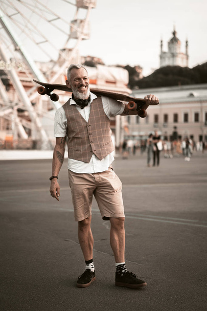 Stylish hipster bearded man with skateboard. Adult model in casual clothes ride longboard on city street. Street lifestyle sport photo - Foto, Imagem