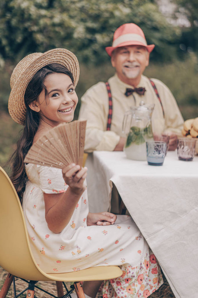 Close up portrait of granddaughter and grandfather at the table in the garden. Happy family relationships. Elderly man with a little girl smiling at the camera - Photo, Image