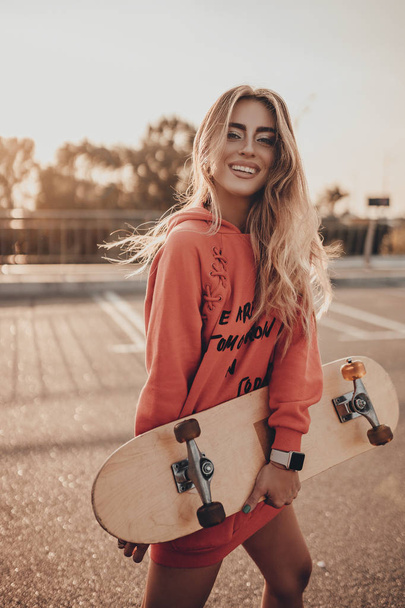 Positive girl walking on city street with skateboard in urban red hoodie. Happy attractive young woman posing on sunset in summer on parking - Фото, изображение