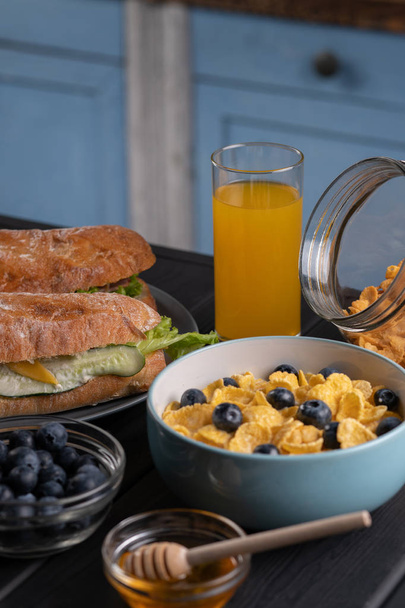 Homemade breakfast. Traditional food. Sandwich, baguette, corn flakes. With sweet berry, honey, muffin. Kitchen. Black table - Фото, изображение