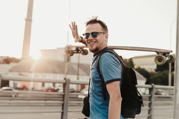Gelukkige hipster die poseert met skateboard bij zonsondergang in de zomeravond. Portret van knappe stijlvolle man model in blauw shirt, zonnebril en hoed op straat achtergrond - Foto, afbeelding
