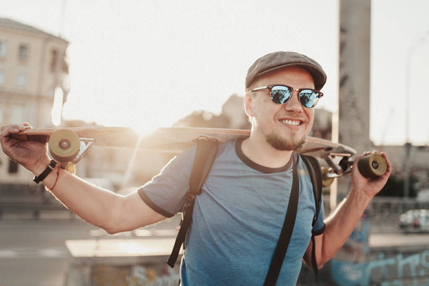 Happy hipster guy posing with skateboard in sunset in summer evening. Portrait of handsome stylish man model in blue shirt, sunglasses and hat on city street background - Zdjęcie, obraz